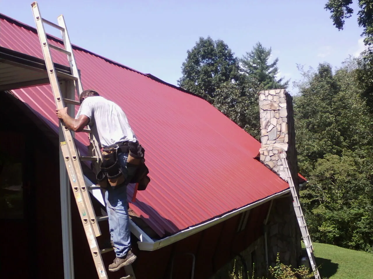 Roofer on a ladder beside a red metal roof and stone chimney — Soffit & Fascia Repair by Roofing Brothers in Barhamsville